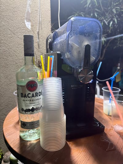 The image displays a nighttime setup for preparing and serving frozen alcoholic beverages, likely on a patio or outdoor area in Kochav Yair Tzur Yigal, Israel.

The central object is a black slushie machine with a clear top reservoir filled with a white, icy mixture, indicating active use. A silver knob and dispensing lever are prominent on the machine, which is labeled "SLUSHYMAKER" and also shows "D51" on a green tag.

On a wooden table in the foreground, an almost empty clear glass bottle of "BACARDÍ CARTA BLANCA SUPERIOR WHITE RUM" rests on its side. Beside it, a stack of clear plastic cups is ready for serving. A collection of colorful plastic straws in blue, yellow, orange, and pink are visible, some placed loosely near the cups.

In the background, two partially filled clear plastic cups with pink straws suggest that drinks have already been served or are in preparation. One of these cups also has a blue straw tied into a knot. The scene is illuminated by artificial light, creating highlights and shadows against a dark background with blurred green foliage, consistent with an evening outdoor gathering.