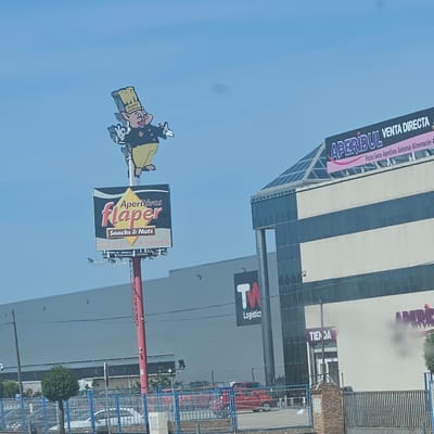 The image shows a roadside advertisement in Azuqueca de Henares, Spain. Prominently displayed is the "Flaper" logo for a snack and nut company, featuring a cartoon pig chef. The sign advertises "Aperitivos Flaper" and "Snacks & Nuts," and includes a website and telephone number.

In the background, a large industrial building displays "TM Logistics" signage. To the right, another building is visible with the "Aperidul Venta Directa" sign, listing products like nuts, snacks, and food items. The word "Tienda" (store) is visible by an entrance. A blue fence borders the road, with various vehicles parked nearby. A small, round tree is planted along the fence line. The sky is clear and blue.