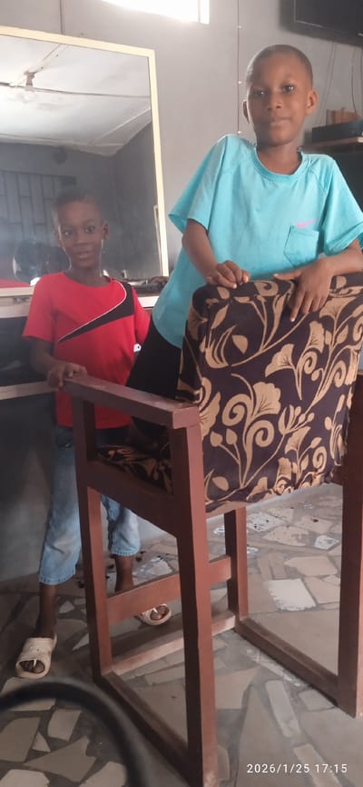 Two young boys pose beside and behind a wooden chair with a brown floral cushion. The boy on the left wears a red shirt with black accents and jeans, while the boy on the right sports a light blue t-shirt. Both are wearing sandals. A mirror is visible behind them. The room appears to be indoors, possibly a shop, with a tiled floor. A date stamp, "2026/1/25 17:15," is visible in the bottom right corner of the image.