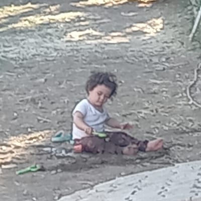 A young child with curly dark hair sits on a gravelly surface, wearing a white t-shirt and dark pants. The child is holding a small green tool, possibly a toy shovel, and appears to be interacting with a pile of dirt or sand. To the left of the child, a pair of green shoes are visible. The background consists of more gravel and some patches of light-colored material. The scene suggests an outdoor setting, possibly a playground or an undeveloped area within Çatalca, Turkey.