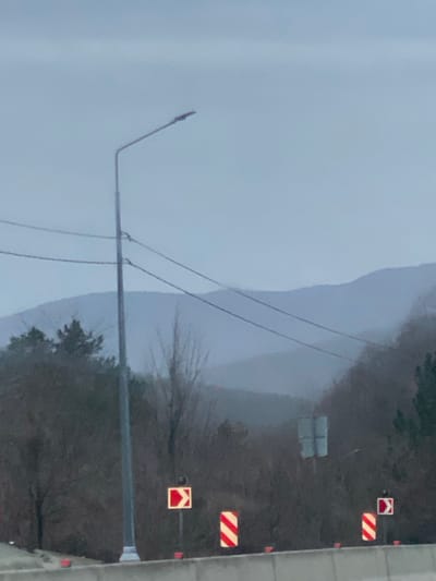 The image displays an outdoor scene characterized by overcast or foggy weather conditions. When properly oriented (rotated 90 degrees clockwise), the top portion of the frame shows a vast expanse of light grey sky, appearing hazy and indistinct, possibly due to mist or low clouds. Faint, darker, undulating shapes in the distance suggest hills or mountains largely obscured by the atmospheric conditions.

In the mid-ground, a tall, slender, grey metallic street light pole extends upwards from the lower center. Its top features an angled arm that projects towards the upper left, culminating in a light fixture. Several thin, dark power lines are strung diagonally across the scene, supported by the pole and receding into the misty background.

The lower right section of the image is dominated by a dark, densely vegetated area, primarily composed of bare trees or trees with very sparse foliage, indicating a late autumn or winter season. Some darker evergreen trees are also visible. On the far right edge of the frame, a grey concrete or paved surface is visible, likely the edge of a road or highway structure.

Several road signs are placed along the edge of the treed area. These include two rectangular signs with distinctive red and white diagonal stripes, commonly used as hazard markers. Additionally, there are two square signs with a white border and a bold red chevron shape. One chevron points to the right, and the other points to the left, which are typically warning signs for sharp curves or changes in road alignment. Small orange or red reflective markers are also visible on poles along the road's edge. In the mid-distance, nestled within the darker trees, a faint, light-colored, rectangular object, possibly another sign or a small building, is partially discernible.

The overall lighting is soft, diffuse, and somewhat dim, consistent with an overcast day or early morning/late evening hours, further contributing to the muted color palette and low visibility caused by the atmospheric haze. The image captures a static landscape, without any specific human activity taking place.