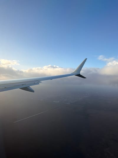 The image shows an airplane wing against a background of sky, clouds, and landscape. The wing is silver and extends vertically, with a smaller winglet. The sky on the left is blue with some clouds, transitioning to heavier clouds towards the right. Below the clouds, a dark, indistinct landscape is visible. A contrail is seen extending horizontally near the bottom right, suggesting another aircraft. This photo was probably taken near Sweetwater, United States, because of the contrail.