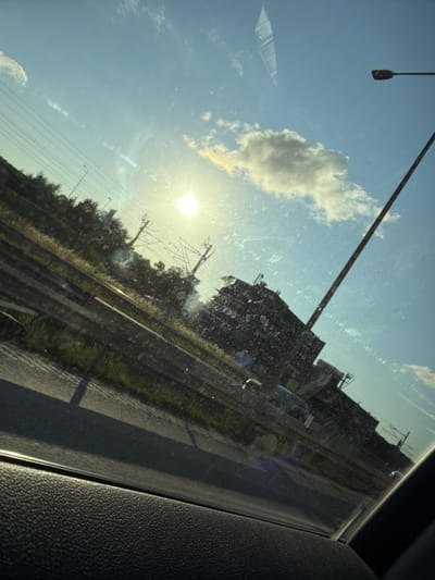 The image shows a view from inside a vehicle, looking out through a window, likely a windshield or side window, at an urban or suburban landscape. The sun is low in the sky, creating a bright glare and lens flare, with visible dust or smudges on the glass.
In the background, a large, dark, multi-story building with visible structural elements and windows dominates the scene. To the left of the building, there are trees and vegetation. Overhead, there are power lines supported by utility poles, and a tall, thin mast-like structure, possibly a lamppost or antenna, extends upwards towards the sky. The sky is blue with scattered white clouds.
A road or highway is visible in the foreground, with a concrete barrier separating it from the embankment. The embankment has overgrown grass and some dry vegetation. There are a few vehicles visible on the road in the distance. The bottom of the image shows the interior of the vehicle, specifically the textured dashboard or door panel.
The overall impression is of a journey through a developed area during daylight, with the sun creating a somewhat hazy and reflective effect. The building in the background suggests an industrial or commercial district.
While the image depicts an urban environment, it is not possible to definitively identify the specific location within Mölndal, Sweden, based on the visual information provided. The presence of overhead power lines, a large building, and a highway are common features in many developed areas.
