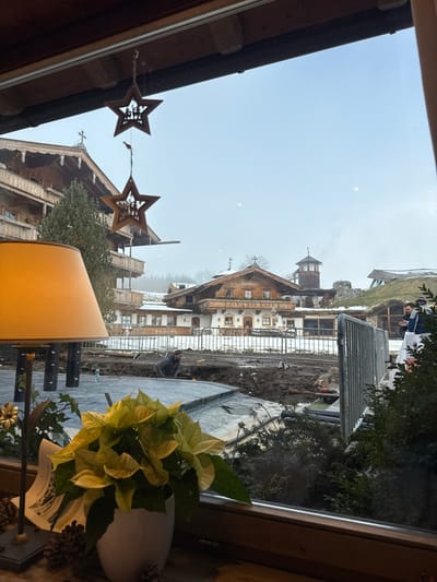 The image captures a view from inside a building in Going am Wilden Kaiser, Austria, looking out through a window onto a winter outdoor scene. In the immediate foreground, a wooden window frame borders the view. On the left, a table lamp with an orange-brown shade is partially visible, alongside a white potted plant with light green leaves and several pinecones resting on the windowsill. Two wooden, star-shaped decorations, each featuring a cutout nativity scene, hang from the upper window frame.
Outside, a construction site is present directly beyond the window. The ground is muddy and excavated, showing a trench with exposed pipes. Silver metal barricades enclose portions of this construction area. A man wearing dark clothing is observed kneeling within the trench on the left side of the frame, appearing to work. To the right, partially obscured by a metal barrier and green foliage, two individuals in dark attire are standing.
In the background, several traditional multi-story alpine-style buildings are visible, characterized by wooden balconies, gabled roofs, and decorative elements. Patches of snow are visible on the roofs. A central building prominently displays the name "Stanglwirt" on its facade. Further to the right, a building with a distinctive green, grass-covered roof and a tower with a conical roof are also present. The distant landscape consists of snow-dusted hills and sparse trees under an overcast or hazy sky.