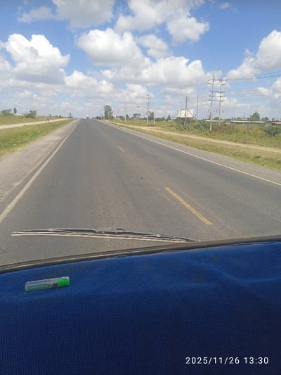 The image depicts a straight, two-lane asphalt road extending into the distance under a blue sky with numerous cumulus clouds. The perspective is from inside a vehicle, looking out through the windshield, with the date and time "2025/11/26 13:30" visible in the lower right.
The road features a dashed yellow center line and solid white lines marking the shoulders. On the immediate left, a dirt shoulder and grassy verge transition to an unpaved track or secondary road. Further left, scattered trees and utility poles are visible, along with a lone individual walking eastward in the mid-distance. On the immediate right, a dirt shoulder and grassy verge lead to a landscape populated by multiple utility poles with overhead power lines. In the mid-distance on the right, several large, cylindrical white tanks on elevated platforms, likely storage tanks or silos, are present alongside other industrial or agricultural structures.
In the immediate foreground, a dark blue, textured fabric covers the vehicle dashboard. A single windshield wiper blade is visible at the base of the windshield. A transparent cylindrical object with a green cap, resembling a pen or a lighter, rests horizontally on the blue dashboard fabric. The scene is located in Kaputiei North ward, Kenya.