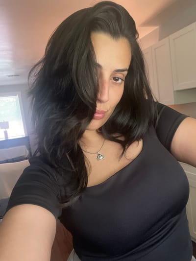 A woman with dark hair and a black top is pictured indoors, with a portion of a room visible in the background. The room appears to be a kitchen or dining area, with white cabinets and a countertop. There is also a window with blinds, and a lamp in the background. The woman is looking at the camera with her hair partially covering her face. She is wearing a silver necklace with a crab pendant. The image is taken from a medium close-up angle. The location of North Charleston, United States of America is not evident from the image itself.