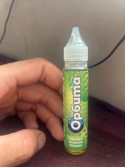 A person's hand is holding a small, clear plastic bottle with a white cap and a green label. The label features the text "Orbita" in large, blue, stylized letters, with the number "80" next to it. Below "Orbita", there is text in Russian that reads "ЖВАЧКА ЗЕЛЕНОЕ ЯБЛОКО" (chewing gum green apple). The bottle contains a yellow liquid. The background is blurred and appears to be a textured surface, possibly a blanket or fabric, in a dark reddish-purple color. The image is taken indoors, and there are no visible signs or features indicating the specific location in the city of Suponevo, Russia.