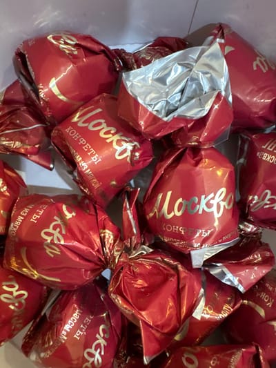 The image shows a close-up, overhead view of a pile of red candy wrappers with gold lettering. The wrappers are crinkled and some appear to be opened, revealing silver foil inside. The lettering on the wrappers appears to be in Russian and includes the word "Москва" (Moscow) and "КОНФЕТЫ" (candies). The context of Chuguyevka, Russia, is not visually represented in the image itself, as it is an indoor shot of a collection of candies. There are no people, actions, or interactions depicted. The environment appears to be a simple, possibly domestic setting, with the candies likely contained within a box or bag, suggested by the light-colored background.