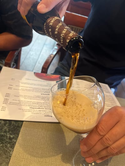 In an indoor commercial setting located in Baarle-Nassau, Netherlands, a close-up perspective captures a person's hands pouring a dark amber liquid from a brown glass bottle into a stemmed glass. The bottle's label features repeated text, partially visible as "BRUIN". The liquid creates a thick layer of tan foam within the tilted glass, which is held by the same individual. On the table, a beige woven placemat and a white menu with Dutch text, including "Soepen" (soups) and "Warme voorgerechten" (warm appetizers) alongside pricing, are present. Background elements include a dark-clothed figure and blurred wooden furniture.