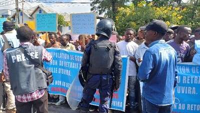 The scene is depicted in what is stated to be Kabinzi, Rwanda, during daylight hours. A crowded outdoor gathering or demonstration is underway on what appears to be a street or open area.

In the foreground, from left to right:
1.  A person, identified as "PRESSE" by a dark grey vest worn over a red and white checkered shirt, is seen from behind. They are holding a device, possibly a smartphone or camera, up to eye level, seemingly documenting the event. A dark bag strap crosses their shoulder.
2.  A riot police officer, viewed from behind, stands centrally. The officer wears a dark blue uniform, black body armor for the torso and arms, and a black helmet. They hold a clear, rectangular riot shield, resting its bottom edge on the ground to their left.
3.  A third individual is seen from behind and slightly to the right, wearing a blue denim shirt and a black baseball cap. They are looking towards the crowd and the officer.

The background features a large group of individuals, predominantly male, forming a crowd behind the police officer and extending to the sides. Several members of the crowd are holding light blue and yellow placards with dark writing, which is largely illegible. A large blue banner, partially visible, is held horizontally by multiple people in the mid-ground. Text on the banner includes "POPULATION DE GOM...", "NON AU RETRAIT...", and "ILLE D'UVIRA." and "BURUNDAI", indicating a protest related to the withdrawal from Uvira, a city in the Democratic Republic of Congo.

Behind the crowd, several residential-style buildings with light-colored walls and dark roofs are visible. Green trees and yellow flowering bushes are present in the upper right background under a bright, clear sky.