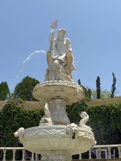 A low-angle shot depicts a water fountain in Tehuixtla, Mexico, showing a classical statue of a male figure holding a trident. The sculpture is rendered in white stone, and water flows from its base into a tiered fountain structure below. This fountain section includes sculpted lion heads and floral motifs. Behind the fountain is a backdrop of lush green foliage, including trimmed hedges and tall, slender trees, suggesting a formal garden setting. The sky is clear and blue, indicating a sunny day. A portion of a white balustrade fence is visible to the right of the fountain.