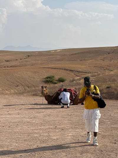 The image captures a scene in Sidi Badhaj, Morocco. A man wearing a yellow shirt, white pants, and sneakers stands near a camel. He has a black bag and is looking at a mobile phone in his hands. Another man, dressed in white, is kneeling by the camel. The camel has a saddle covered with a red and white pattern. The ground is sandy, and the background shows a dry, hilly landscape under a partly cloudy sky.