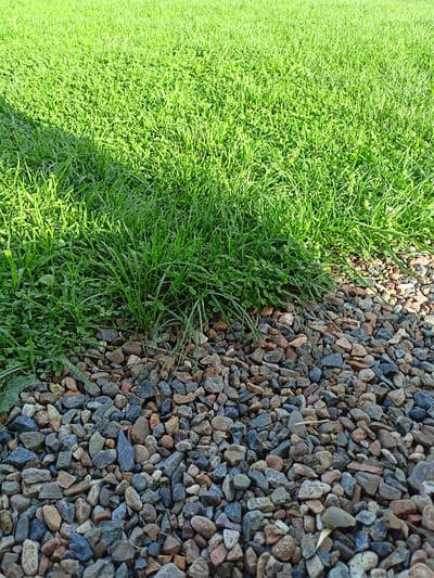 The image shows a close-up view of a patch of green grass meeting a border of small, multi-colored stones. A shadow from an unseen object falls across the upper left portion of the grass. The scene appears to be an outdoor area in a park or garden, possibly in Kurtukovo, Russia, though this location cannot be definitively confirmed from the visual information alone. There are no people or animals visible in the image, nor are there any discernible actions or interactions taking place. The environment is characterized by the contrast between the soft texture of the grass and the rough texture of the stones.