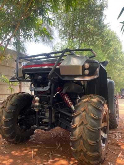 The image, taken in Dhundshi, India, presents a robust, grey and black All-Terrain Vehicle (ATV) or Utility Task Vehicle (UTV) parked outdoors. The vehicle is oriented vertically, suggesting the photo was taken with the camera rotated 90 degrees clockwise.
The ATV features large, aggressive-tread tires designed for off-road use, fitted with bright, possibly chrome or silver-colored rims. Visible components include red coil springs on the suspension, suggesting heavy-duty shock absorption, and a black exhaust pipe. The front of the vehicle is equipped with a sturdy black roll cage or bumper and two round LED headlights. A tow hitch with a chrome ball is visible towards the rear underside. The side of the ATV bears the visible text "POWERLAND" in white letters with a red accent line.
The setting appears to be a natural, unpaved environment. The ground beneath the ATV is covered in reddish-brown dirt or soil, with scattered dry leaves and debris. Lush green foliage, including bamboo plants and other leafy trees, frames the vehicle, with the bright sky visible through the canopy. The clear, bright sunlight indicates it is daytime, casting subtle shadows. No people or activities are visible; the ATV is stationary.