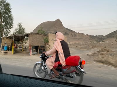 The image shows a man wearing a pink headscarf, driving a red motorcycle along a paved road near a small town in the Panjwai district of Kandahar Province, Afghanistan, located at latitude: 31.616, longitude: 65.591. The motorcycle has a large, red bag attached to the back. The man is wearing a dark-colored long-sleeved shirt underneath the scarf. The image is taken from the passenger seat of a car that is traveling behind the motorcycle. In the background, the road passes by a small town with a few simple structures made of mud bricks and straw. There is a hill in the background, and a vast, barren landscape stretches out in front of the motorcycle. The sky is clear and blue. The image has a slight blur due to motion.  The scene is typical of the arid landscape of southwestern Afghanistan, featuring the mountains, plains, and small towns.