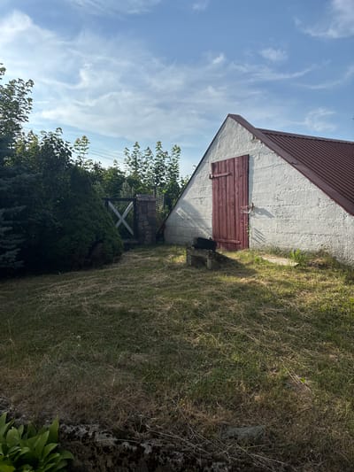 The image shows an outdoor scene with a shed, a gate, and vegetation under a partly cloudy sky. The shed is white with a pitched, dark red metal roof. It has a wooden door painted dark red with vertical planks and metal hinges and latch. To the left of the shed, there is a gate made of stone pillars and a wooden cross-shaped structure. Dense green bushes and trees are visible to the left of the gate. The ground is covered with dry grass and some green patches. A small, dark object, possibly a tire, rests on a concrete block near the shed. The overall setting appears to be a rural or suburban area. The provided information states the location is Janów, Poland. There are no people visible in the image.
