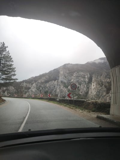 The view from inside a vehicle as it enters a tunnel. The scene is set on a winding road in Lyutibrod, Bulgaria, with a mountain range in the background. The range is predominantly composed of light-colored, rugged rock formations. The foliage is sparse, with trees and shrubs clinging to the slopes. The weather is overcast, and the mountaintops are shrouded in mist or low clouds. A 40 km/h speed limit sign is visible. The road is marked with a solid white line, and chevron signs line the roadside indicating the curve of the road. A single tree stands beside the road. The interior of the vehicle, specifically the dashboard, is visible in the foreground.