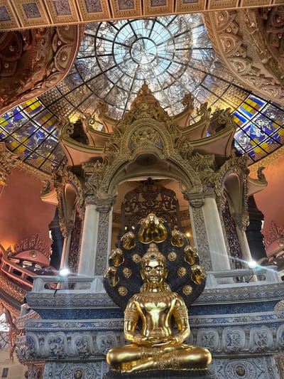 The image depicts an ornate interior space in Samrong Nuea Subdistrict Municipality, Thailand, featuring a golden reclining Buddha statue. Above the statue, there's a decorative ceiling with a glass dome displaying map-like designs and stained-glass windows with blue, yellow, and white elements. The architecture includes intricate carvings and moldings with gold accents. The pillars and beams feature detailed patterns and designs.