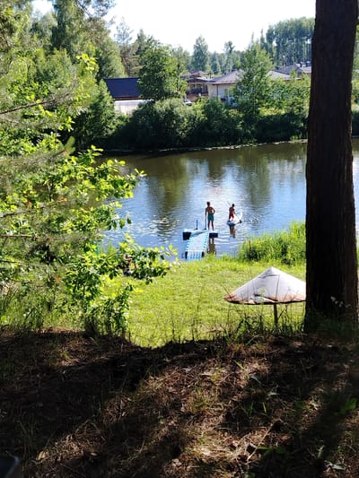 The image depicts a serene summer scene at Stroitel', Russia. Two individuals are engaged in paddleboarding on a calm body of water, likely a river or lake, surrounded by lush green trees and vegetation. In the foreground, a large tree trunk partially obscures the view, and a rusted, umbrella-like object rests on the grassy bank. The water reflects the clear blue sky, suggesting fair weather and daytime. A blue floating dock is visible near the shore, with one person standing on it and the other on a paddleboard. The surrounding area appears to be a residential zone, with houses nestled among the trees in the background, indicating a suburban or semi-rural setting. The overall atmosphere is peaceful and recreational, highlighting a pleasant summer day spent outdoors.