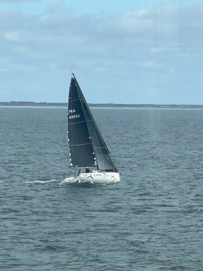A white-hulled sailboat, featuring a black mainsail marked "FRA 43933" and white hull branding "V1D2 SL ENERGIES," is actively sailing through choppy, dark blue-gray waters offshore from Ploemeur, France. The vessel generates a white wake behind its stern, indicating forward motion. A single individual, partially visible in a light-colored jacket, is present in the cockpit area. A low, distant coastline is discernible on the horizon beneath a light blue sky with scattered white clouds.