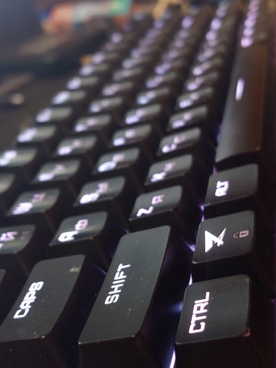 The image is a close-up, angled shot of a black keyboard with illuminated keys. The lighting is dim, with purple and white light emanating from the keys, suggesting a gaming or custom keyboard setup. The keys visible include "CAPS", "SHIFT", "CTRL", "ALT", and several other alphanumeric and symbol keys. The texture of the keys appears to be matte plastic. The background is out of focus and appears to be a desk or table surface, with blurred colors suggesting other electronic devices or a dimly lit room. The image is a still photograph and does not depict any people or actions. There is no discernible information in the image that would indicate its location in Sokolohirsk, Ukraine.