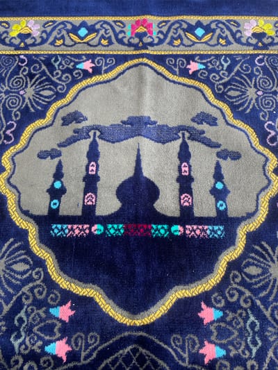 The image shows a close-up, overhead view of a navy blue prayer rug with intricate designs. The central motif features a silhouette of mosque minarets against a grey background, outlined by a wavy gold border. The minarets are decorated with pink and blue accents. The rug's outer edges and surrounding area are adorned with swirling grey patterns and small, colorful floral or star-like embellishments in pink, yellow, and blue. The texture of the rug is visible, appearing soft and possibly made of a plush material. The lighting suggests it is indoors. There is no visible indication of the location being Trieng Paloh, Indonesia, or any specific environmental context beyond the rug itself. The image is solely focused on the design and texture of the prayer rug.