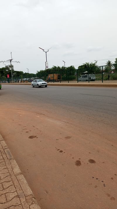 The image depicts a street scene in Kawo, Nigeria. A silver car is driving on a gray asphalt road with a yellow median. To the left is a red-brown pavement with several dark stains. A sidewalk with patterned stone is visible on the lower left. On the other side of the road, a fence separates the street from trees, a van, and a light-colored waste management truck. Utility poles with multiple lights and some signs stand near the background trees. A pedestrian is walking along the road. The sky is overcast.