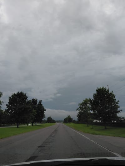 The image depicts a two-lane asphalt road with cracked pavement, possibly indicating wear and tear. The road stretches into the distance, framed by lush green grass and lined with mature trees on both sides. The sky is overcast with heavy, dark clouds, suggesting imminent rain or stormy weather. Power lines are visible against the cloudy sky on the right side of the image. A few vehicles are present on the road, some with their headlights on. The overall scene suggests a rural or suburban setting outside of a major city. Based on the style of the road, vegetation, and architecture visible in the background, this scene could be located in Lexington, Kentucky, United States of America, likely on a road leading out of the city.