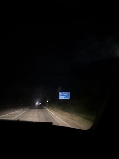 A nighttime, slightly tilted, eye-level view of a highway in Pisanets, Bulgaria, as seen from inside a vehicle. The scene is predominantly dark, with the road illuminated by headlights from oncoming traffic and the vehicle the photo is taken from. The road surface is visible, with white lane markings and a reflective barrier along the right side. A blue highway sign with white text and directional arrows is visible on the left side of the road. The sign appears to indicate directions to "Pisanets" and "General Toshevo" with arrows pointing left and right respectively. The weather appears clear, with no visible precipitation. The overall mood is one of transit and travel in a rural, dark environment.
