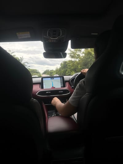 The image captures the interior of a vehicle, viewed from a rear passenger seat, in the city of Khian Sa, Thailand. A driver, identifiable by the back of their head and right arm, is visible in the front left seat. The driver is wearing a light-colored short-sleeved top; their left hand is on the steering wheel, and their right hand rests near the gear shifter on the center console. The vehicle's dashboard and central console are trimmed in a dark red material, accented with silver-colored details around the air vents. A large infotainment screen displays a navigation map with a blue route line, a vehicle icon, and numerical data including "40 km," "120 km/h," and a timestamp of "15:46." Above the dashboard, a rearview mirror and an overhead console are visible, along with an "AIRBAG" warning sticker affixed to the passenger-side sun visor. Through the windshield, an overcast sky with grey clouds is visible above a road flanked by dense green foliage, including palm trees on the left. Several white vehicles are discernible in the distance on the road ahead.