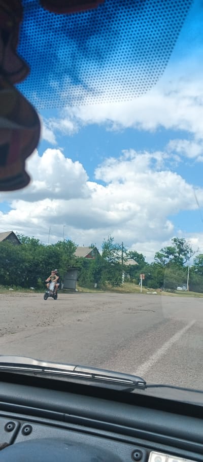 The image, taken from inside a vehicle, captures a scene in Mala Smilianka, Ukraine. The view is through the windshield, which is partially obscured by a dark object at the top left and a pattern of small dots across the blue sky. Two people are riding on a light-colored scooter on a dirt road. The person in front is driving, wearing a dark t-shirt and shorts, while the person behind them is wearing a light-colored t-shirt and shorts. They are surrounded by greenery, including trees and bushes, and several houses with red and white roofs are visible in the background. The sky is predominantly blue with scattered white clouds. A metal fence is located to the left of the scooter. The road surface appears to be a mixture of dirt and asphalt, with some visible potholes. In the distance, a silver car is parked on the right side of the road. The interior of the vehicle, including the dashboard and windshield wipers, is visible in the foreground.