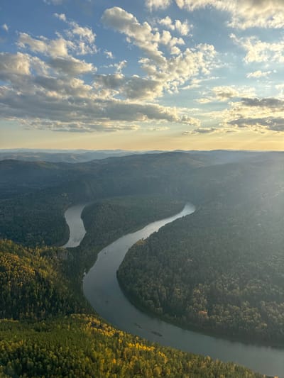 An aerial image captures a vast natural landscape in Manskii, Russia. A wide, serpentine river, appearing gray to silver under indirect light, dominates the middle ground, flowing through a pronounced S-curve. The river is flanked on both sides by extensive, dense forests covering rolling hills and valleys. The forest canopy is primarily dark green, with noticeable patches of lighter green and distinct areas of yellow foliage in the foreground, suggesting autumn conditions. In the distance, multiple layers of hazy blue-gray mountains extend to the horizon. The sky occupies the upper third of the frame, displaying a light blue color with scattered white and gray cumulus clouds. Low-angle sunlight emanates from the right side of the frame, casting a warm, yellowish glow on the horizon and illuminating parts of the clouds. No human structures, vehicles, or individuals are discernible within the scene.