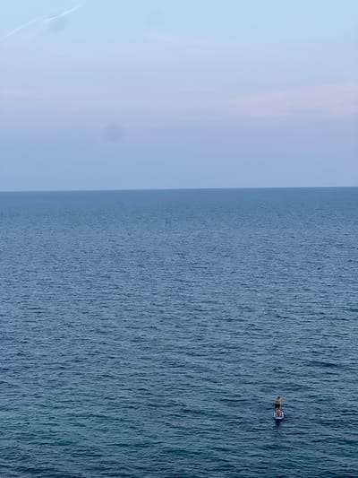 A wide shot captures two individuals on paddleboards in the calm, blue waters of the sea, likely off the coast of Ovingdean, United Kingdom. The horizon line is clear, separating the vast expanse of the ocean from a pale blue sky with wispy clouds. The water is slightly rippled, with a gradient of blues, transitioning from a darker shade in the foreground to a lighter hue near the horizon. One paddleboarder stands, appearing to paddle, while the other is seated on their board. The scene conveys a sense of tranquility and outdoor activity.