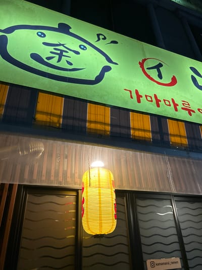 An illuminated nighttime exterior view captures the facade of a business, identified as "Kamamaru Ramen" (가마마루 라멘), located in 서대문구, South Korea. The upper portion of the image features a large, bright green illuminated sign. On the left side of the sign, a stylized black outline of a pot with blue eyes and the Chinese character "余" is depicted. To the right, the Japanese katakana character "ス" (su) is rendered in red, followed by the Korean text "가마마루" also in red.
Below the main sign, a horizontal section of the building displays alternating dark blue/black and yellow/orange vertical stripes, possibly an awning or decorative panel. Below this, a translucent, corrugated sheet material extends across the width, showing a faint reflection of an internal or overhead structure. Centrally positioned in the lower half of the image, a large, cylindrical yellow paper lantern hangs, glowing brightly and featuring subtle red patterns on its ribbed surface. Below the translucent panel and to the right of the lantern, dark-framed windows with wavy, frosted patterns are visible. In the bottom right corner, a small white sign with black text displays "@kamamarui_ramen", indicating an Instagram handle. No individuals are present in the image.