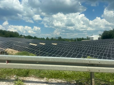 A solar farm is situated in a rural, hilly area under a partly cloudy sky in Vilusi, Montenegro. The foreground features a metal guardrail, common along roadsides, with green grass and some sparse vegetation growing beneath it. Beyond the guardrail, a vast expanse of dark solar panels stretches across the landscape, arranged in neat rows. In the distance, rolling hills covered in trees are visible, and a white building with a modern, angular roof stands among the trees. The sky is a mix of blue and white clouds, suggesting fair weather.
