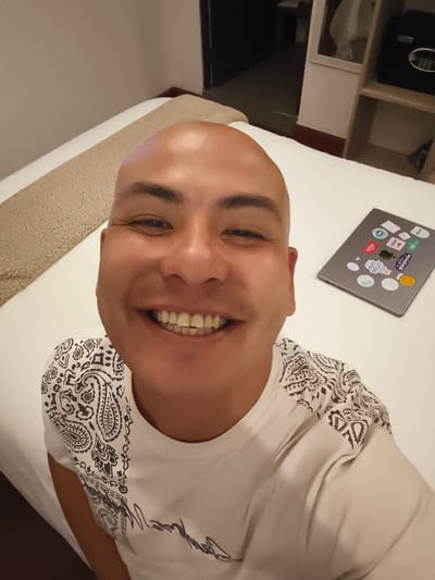 The image shows a bald man smiling broadly, with a close-up perspective. He is wearing a white t-shirt with black paisley patterns on the sleeves. He appears to be lying on a white bed, next to a laptop covered in stickers. Behind him is a headboard, a bedside table, and part of a wall. The photograph was possibly taken in Guatemala City, Guatemala.
