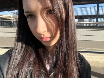 The image depicts a close-up portrait of a young adult female, positioned outdoors on what appears to be a train or metro platform in Alfortville, France. The female has long, straight, dark brown hair that falls over her shoulders, and light brown eyes. Her face is unevenly illuminated by strong sunlight, casting distinct linear shadows across her left eye, nose, and forehead. She is wearing a black jacket, consistent with leather or a similar material, over a light-colored, possibly knitted, top. A silver-toned chain is visible around her neck. Her expression is neutral, with her lips slightly parted. The background, largely out of focus, reveals grey concrete platforms, a metal fence or railing, and overhead electrical lines or support structures against a clear blue sky. Buildings are faintly visible in the far distance, suggesting an urban or suburban setting.
