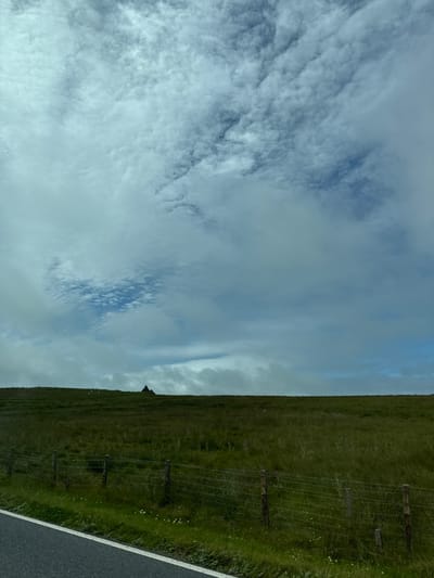 A wide shot depicts a cloudy sky over a green, grassy hill. The horizon line is low, with the sky occupying approximately two-thirds of the frame. Wispy, textured clouds are scattered across the sky, with patches of blue visible. On the distant hill, a small, dark, triangular structure, possibly a building or a shed, is visible. A wire fence runs across the foreground, parallel to a paved road on the left side of the image. The road has a white line marking its edge. Sheep are visible as small white specks on the grassy hill to the left of the structure. The overall impression is a rural landscape under a dynamic sky.