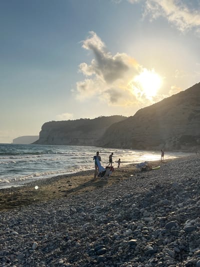 The image depicts a sunlit beach scene with a rocky shore and a calm sea. The sun is low in the sky, casting a warm glow and creating a bright highlight on the water's surface. The sky is partly cloudy, with some fluffy white clouds and larger, darker ones near the sun, indicating it might be late afternoon or early morning.

Several people are visible on the beach, engaged in various activities. Some are walking along the water's edge, while others appear to be sitting or resting. One person stands near the water, looking out towards the sea, while another group is gathered further up the beach. There's a hint of a cliff or rock formation in the background, suggesting a natural, somewhat rugged coastal environment. The beach is covered in smooth, rounded pebbles of various grey and brown tones. The water is gently lapping at the shore, creating small waves and reflecting the sunlight.

The overall mood is peaceful and relaxed, characteristic of a day spent at the seaside. The lighting suggests a pleasant, possibly warm, weather condition. The presence of people enjoying the beach implies a recreational or leisure setting. The scene captures a serene moment by the ocean. No visible text is present in the image.