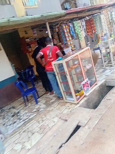 The image, taken from a high-angle perspective looking down into a retail space, depicts a scene within what appears to be a market stall or small shop.
In the mid-ground, a person wearing a red short-sleeved t-shirt and dark trousers (possibly jeans) is standing with their back mostly towards the camera. They have a dark strap or bag slung across their left shoulder. Their head, covered in dark hair, is slightly bent downwards as if attending to something on a surface in front of them. To their right, largely obscured by their body, another person in dark clothing is seated behind what appears to be a counter or table.
To the immediate right of the standing person, there is a glass display case with a light wooden frame. Inside the case, some indistinct items are visible, possibly food or small merchandise. On top of this display case, a small red and white label or sticker is affixed, which might be a QR code or a brand tag, but its text is illegible. Further to the right and slightly behind, two bright blue plastic chairs are placed on the tiled floor.
The left side of the scene (relative to the standing person) features multiple rows of shelving packed with various colorful products. These appear to be packaged goods like snacks, candies, or other retail items, displaying patterns in colors such as orange, red, green, blue, and yellow. The shelving structure appears to be made of metal. The background behind these shelves shows the interior of the shop, with some darker areas and possibly additional storage.
The right side of the image reveals a textured wall, possibly made of patterned tiles or stone in shades of beige and brown. A dark, recessed area or opening is visible in this wall, suggesting a structural feature or another part of the building. The floor throughout the visible area is covered with light-colored, patterned tiles.
The lighting suggests it is either daytime or there is ample artificial illumination. The overall activity appears to be the regular operation of a retail establishment. No clearly legible text is visible other than the faint, unidentifiable label on the display case.