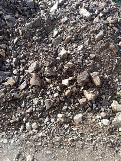 The image depicts a close-up, top-down view of a pile of gravel and rocks. The material ranges in size from small pebbles to larger stones, with colors varying from light gray and tan to dark brown and reddish hues. The rocks appear to be a mix of angular and somewhat rounded fragments, suggesting they may have been quarried or naturally weathered. The ground in the foreground is a mixture of smaller gravel and dirt. The scene is outdoors, under natural lighting, with shadows cast by some of the larger rocks indicating the presence of sunlight. There are no people or any other identifiable subjects present in the image. The context suggests an outdoor location, likely a construction site, quarry, or roadside where aggregate materials are stored. The image does not provide any specific visual cues that would allow for the determination of its location in Bykov, Russia.