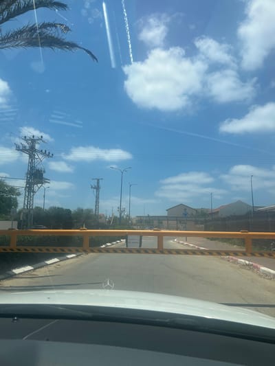 The image shows a barrier gate across a road, likely in Mikhmoret, Israel. The gate is yellow with black and white stripes and appears to be closed. A sign is visible on the right side of the gate with Hebrew text and a phone number, "052-9406941". Beyond the gate, there is a paved road with a white and red striped curb. In the background, there are buildings, trees, and power lines under a blue sky with white clouds. The photo is taken from inside a vehicle, with the hood and windshield of a white car visible in the foreground, showing a Mercedes-Benz emblem on the hood.