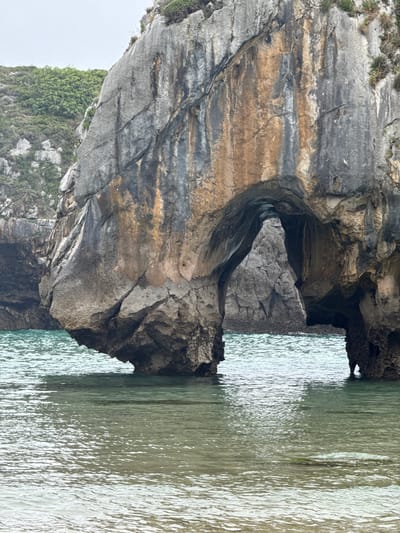 The image shows a natural rock archway carved into a cliff face at the coast. The arch is massive, with rough, weathered rock formations. Through the arch, another smaller rock formation is visible. The water in front of the arch is clear and shallow, with ripples on the surface reflecting the overcast sky. The cliff face is covered in patches of green vegetation. The scene is likely part of the dramatic coastline near Llanes, Spain, known for its natural arches and rugged cliffs. There are no people or man-made objects visible in the image.