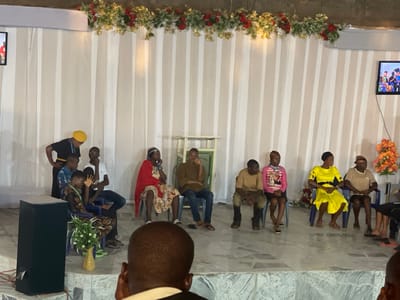 The scene takes place in Nteje, Nigeria. There is a group of people on stage in front of a backdrop of white curtains. Garlands of greenery and red flowers adorn the top of the curtains.
To the left, a man in a yellow cap leans over a seated group of four young adults in chairs. To the right, a group of six individuals sit in chairs. Most are looking down or sideways. A small table is on stage near the center.
In the foreground, the back of a person's head is visible. A speaker and a floral arrangement stand to the side of the stage. A screen can be seen on the right side of the stage.