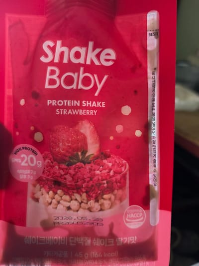 The image shows a pink pouch of "Shake Baby Protein Shake Strawberry" located in Anseong-si, South Korea. The pouch features large white text displaying "Shake Baby" and smaller text indicating "PROTEIN SHAKE STRAWBERRY". A circular emblem on the left side highlights "HIGH PROTEIN" with "20g" and nutritional information in Korean. The pouch also includes a vertical strip on the right with Korean text and the word "BEST!". The background is primarily red with some blurry dark spots. A serving suggestion showing a bowl of the shake with strawberries and granola is depicted on the lower half of the pouch.