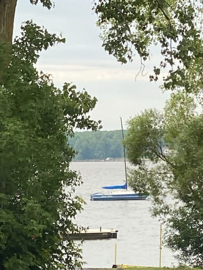 The image shows a serene view of a body of water, likely a lake or river, framed by lush green trees in the foreground and background. In the middle ground, a sailboat with a blue sail is anchored on the water. To the left, a small dock or pier extends into the water, and a single pole stands vertically nearby. The sky is overcast with a soft, diffused light. The scene evokes a sense of tranquility and natural beauty, possibly a summer or early autumn day. The location is identified as Baie-D'Urfé, Canada.