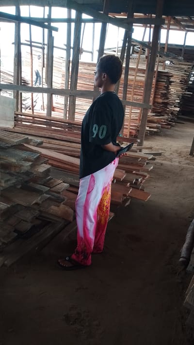 The image shows a woman standing in what appears to be a lumberyard in Asubi, Nigeria. The woman is wearing a black t-shirt with the number "90" on it, along with white and pink pants, and black sandals. She is looking to the right and holding a small black object in her hands. Stacks of lumber are visible around her. In the background, structural beams of the building are visible, and a person can be seen further in the distance behind the beams. The ground appears to be dirt.