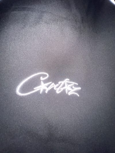 The image shows a close-up of white embroidered text on a dark, possibly black, fabric surface. The text is a cursive script, appearing to spell "Cailla". The background fabric has a visible weave pattern. The location in Krasnoyarsk, Russia is not apparent from the image. There are no people visible.