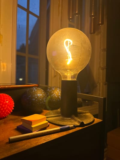A close-up, low-angle shot captures a dimly lit room with a desk in the foreground. The focal point is a vintage-style incandescent lightbulb, its filament glowing warmly, casting a yellow light. To the left of the bulb are three stacks of colorful sticky notes, purple, yellow, and orange, along with a silver pen. Behind the sticky notes are two dark-colored, textured spheres and a vibrant red spiky ball. The background reveals a window with a grid pattern, reflecting a dark blue sky with hints of clouds. The room appears to be situated in an urban environment, possibly an apartment or house in McMasterville, Canada.