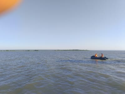 An inflatable boat with two occupants wearing orange life vests is traveling across a body of water under a clear, pale blue sky. The boat appears to be moving from left to right in the frame. One occupant in the front is seated, facing forward, while the other, in the back, is seated and facing left, holding a fishing rod. In the distance, a treeline is visible along the horizon, suggesting land. The water is rippled with moderate waves. An out-of-focus orange blur is present in the upper left corner of the image. The scene is consistent with the Volga River near Krasnyi Yar, Russia, known for its vast expanses of water and recreational activities.